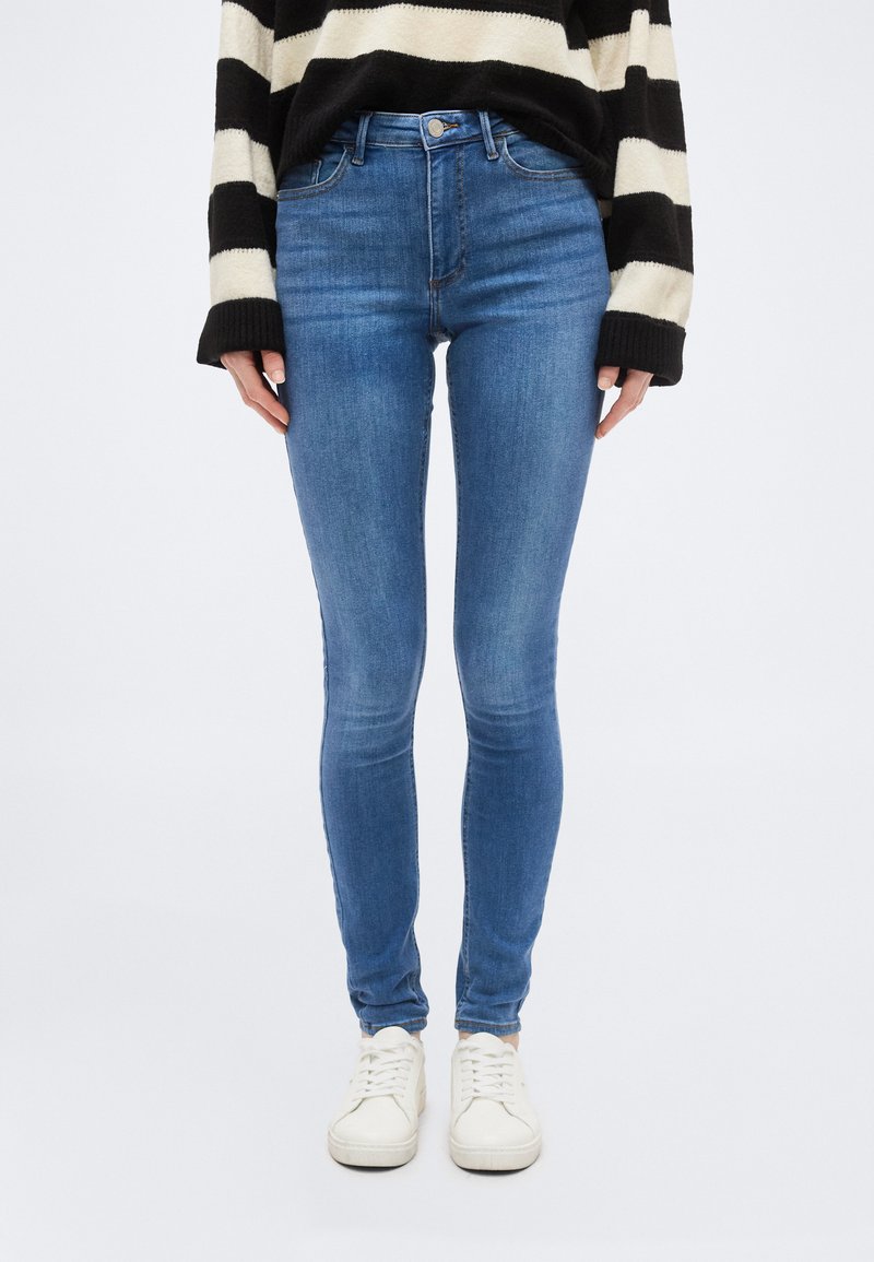 Άτομο που φοράει στενά μπλε τζιν, λευκά αθλητικά παπούτσια και μαύρο-άσπρο μακρυμάνικο πουλόβερ με ρίγες, στέκεται μπροστά σε απλό φόντο.