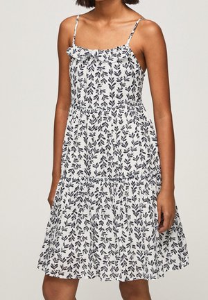 Woman wearing sleeveless white dress with black leafy pattern and ruffled neckline, standing against plain background.