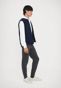 Jauns vīrietis stāv profila stāvoklī, ģērbies baltā kapucē, tumši zila veste, tumšos sporta biksēs, melnās ze Sock uz baltām kedām fona ar vienkāršu fonu.
