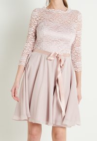 Woman wearing a pale pink dress with lace three-quarter sleeves and a satin ribbon tied at the waist, skirt flaring gently.