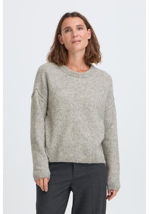 Femme aux cheveux bruns mi-longs, portant un pull ample gris clair et un pantalon gris foncé, debout sur un fond blanc uni.