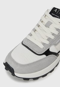 Białe i szare tenisówki z siateczkowego materiału, z zamszowymi nakładkami, czarnymi akcentami, białymi sznurowadłami oraz metką "Armani Exchange" na języku.