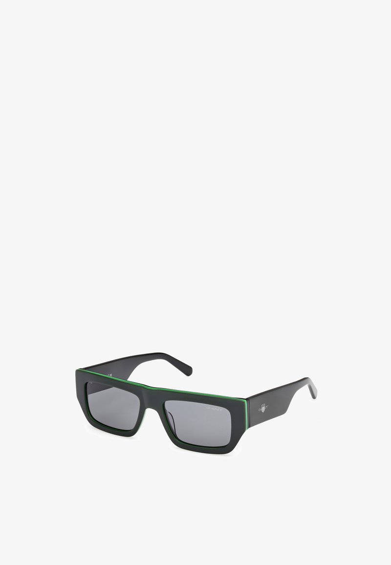 Schwarze Sonnenbrille mit matter Oberfläche und einem grünen Akzent entlang der Oberseite. Quadratische Gläser mit grauem Farbton. Leichter Kunststoffrahmen.