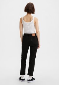 Jeans negros con un diseño de pierna recta, combinados con una camiseta sin mangas blanca. Presentan un parche de cuero marrón en la parte trasera de la cintura y zapatillas de deporte en contraste.