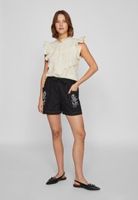 Blusa creme com mangas bufantes e acabamento em renda, combinada com calções pretos com bordados florais e um cós elástico. Sapatos pretos de slip-on.