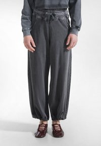 Person trägt lockere graue Hosen mit elastischen Bündchen, gebundener Taille und burgunderrote Schnürflache Schuhe, steht vor weißem Hintergrund.