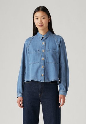 Camicia di jeans cropped in blu chiaro, dotata di chiusura con bottoni, due tasche sul petto e maniche lunghe con polsini elasticizzati.