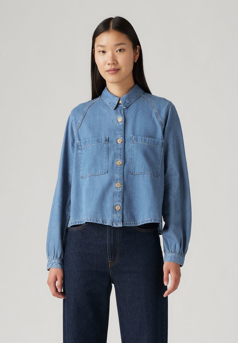 Cropped denim shirt in light blue, featuring a button-up front, two chest pockets, and long sleeves with elastic cuffs.