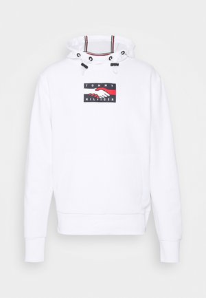 Valkoinen huppari, jossa on etupuolella kengurun tasku, nyörillä varustettu huppu ja grafiikka, jossa on käsiä ja teksti "TOMMY HILFIGER" mustana ja punaisena.