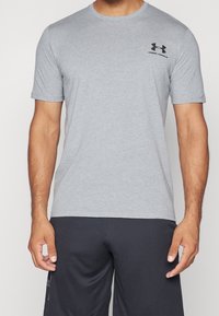 Camiseta atlética de manga corta en gris, hecha de una tela suave, con un cuello redondo y un logo negro de Under Armour en el lado izquierdo del pecho.
