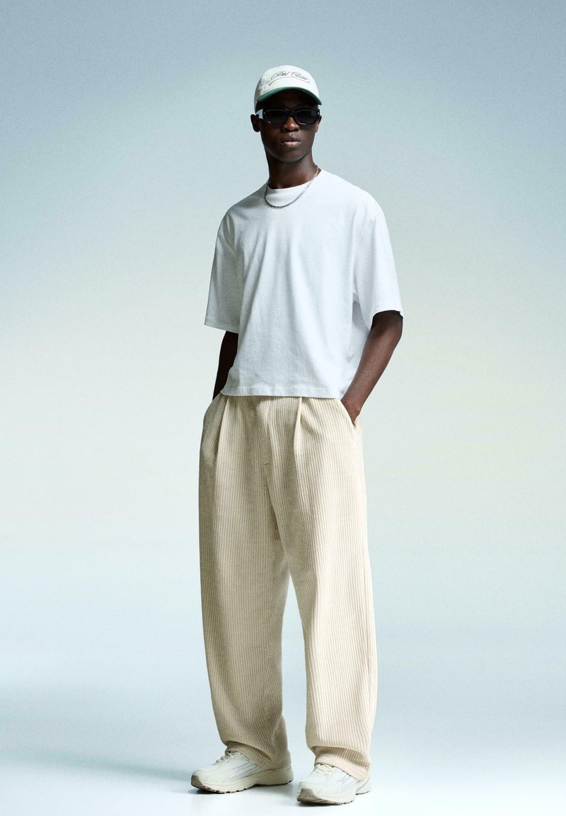 White oversized t-shirt, beige ribbed wide-leg pants, white sneakers, and a light cap. Accessories include dark sunglasses and a necklace.
