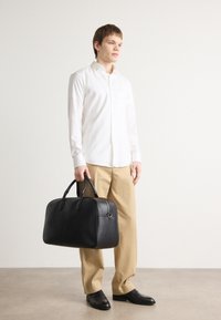 Schwarze Leder-Dufflebag mit doppelten Griffen, rechteckiger Form, glatter Textur und subtilen Hardware-Akzenten. Modell trägt beigefarbene Hose und weißes Hemd.