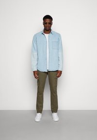 Camicia di jeans azzurra chiara con taschino sul petto, abbinata a pantaloni cargo olive e sneakers bianche, indossata da una persona in piedi dritta.