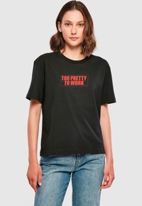 Schwarzes Baumwoll-T-Shirt mit einem rechteckigen roten Aufdruck "TOO PRETTY TO WORK", der auf der Brust zentriert ist. Mit kurzen Ärmeln und rundem Halsausschnitt.