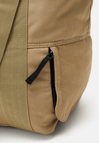 Beige kangaskassi, jossa on teksturoitu verkkomateriaalia sisältävä sivupaneeli, varustettuna pienellä vetoketjullisella taskulla, jossa on musta vetoketjun vetimien korostus.