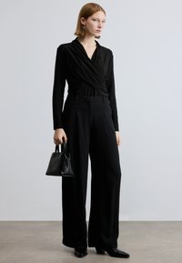 Zwarte lange mouwen overslagtop gecombineerd met hoge taille zwarte wijde broekspijpen. Model houdt een kleine zwarte handtas vast, staand op een neutrale ondergrond.