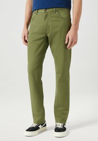 Olivengroene katoenen chinos met een rechte pijp, voorzien van zijzakken, een knoopsluiting, en gedragen met zwart-witte sneakers.