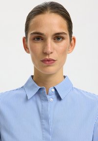 Mujer con el pelo peinado hacia atrás lleva una camisa azul y blanca de rayas finas abotonada, mirando directamente a la cámara con expresión neutral.