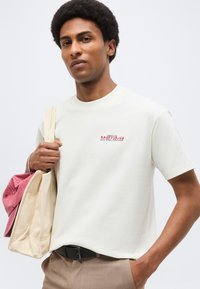 Νέος άνδρας με κοντά σγουρά μαλλιά που φοράει λευκό t-shirt και κρατάει μια μπεζ τσάντα tote με ένα ροζ καπέλο μέσα, στέκεται μπροστά σε ένα απλό φόντο.