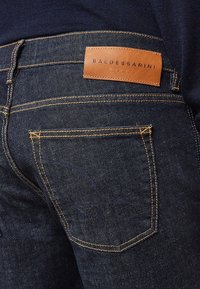 Jeans de mezclilla azul oscuro con costuras naranjas. Parche de cuero en la parte trasera de la cinturilla con el sello "BALDESSARINI". Diseño clásico de cinco bolsillos.