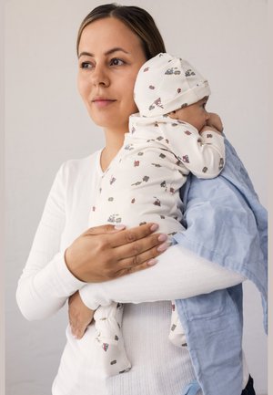 Kvinde i hvidt holder baby iført mønstret heldragt og hat, indpakket i lyseblåt tæppe, kigger til siden mod neutral baggrund.