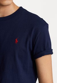 Námořnická modrá bavlněná tričko s krátkými ohrnutými rukávy, s malým červeným logem polo na hrudi. Hladká textura a klasický design s kulatým výstřihem.