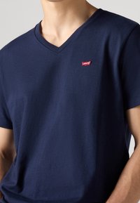 Námořnická modrá bavlněná tričko s výstřihem do V. Má malé červené logo Levi's na levém prsu a krátké rukávy. Hladká textura.