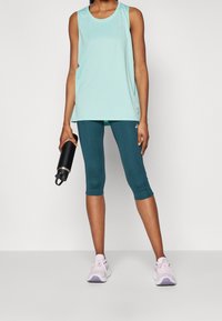 Top sin mangas azul claro, leggings capri en color teal, sosteniendo una botella de agua negra. El calzado son zapatillas deportivas blancas y lavanda.