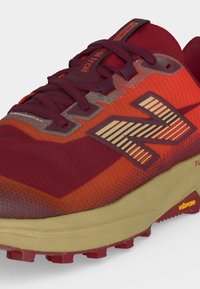Chaussure de trail New Balance rouge et orange avec des lacets bordeaux, une semelle beige et une semelle extérieure Vibram pour une excellente adhérence sur terrain accidenté.