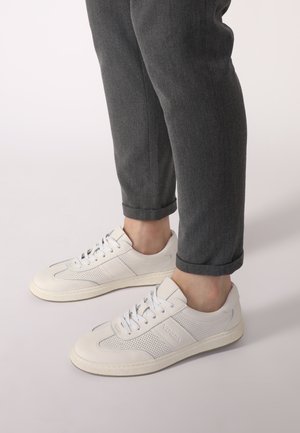 White leather sneakers with perforated panels, textured soles, and gray accents. Match with rolled gray trousers, showcasing a casual style.