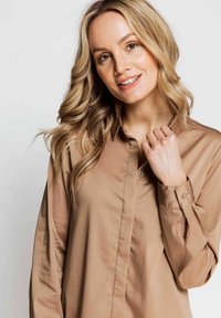 Light brown, long-sleeve shirt with a button-down front, soft texture, and cuffs. Smooth fabric drapes softly, with subtle sheen.