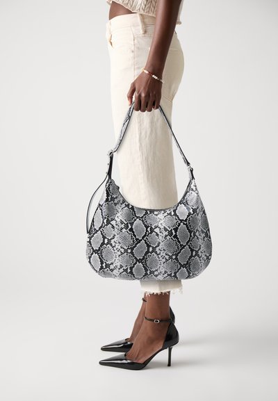Sac hobo en matériau à motif serpent noir et blanc, avec une seule sangle et une texture lisse. Porté avec un pantalon de couleur claire.