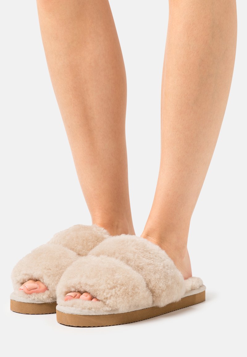 Füße, die beigefarbene, flauschige, offene Pantoffeln mit dicken Sohlen tragen, vor einem schlichten hellen Hintergrund.