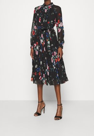 Robe midi noire transparente à imprimé floral multicolore, manches longues, jupe plissée, ceinture nouée à la taille, portée avec des talons hauts noirs à bride cheville.
