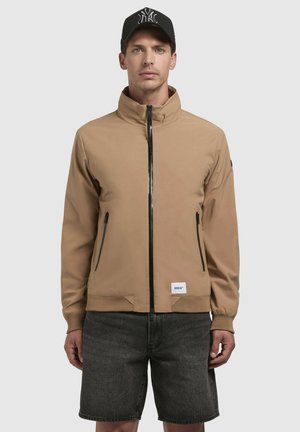 Brun, zip-front jakke med høj krave, to sidelommer og ribbede manchetter. Bæres med grå shorts; har en sort cap med logo.