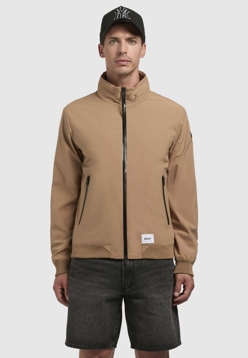 Chaqueta marrón con cremallera frontal y alto cuello, dos bolsillos laterales y puños acanalados. Llevada con pantalones cortos grises; presenta una gorra negra con logotipo.