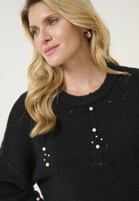 Melna adīta džemperis ar teksturētu rakstu, kam priekšpusē ir dekoratīvas baltas pērles. Apaļa dekoltē un garas piedurknes.