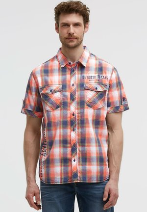Mann trägt ein kurzärmeliges orange, weiß und marine kariertes Hemd mit Brusttaschen und blaue Jeans, steht vor einem einfarbigen Hintergrund.