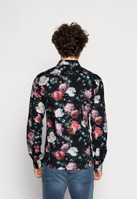 Uomo con capelli ricci che indossa una maglietta nera a maniche lunghe con un motivo floreale colorato, mostrato di spalle su uno sfondo neutro.