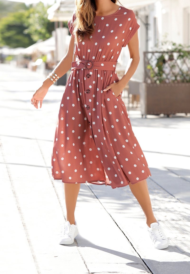 Femme marchant en plein air dans une robe midi couleur rouille à pois blancs, avec ceinture, manches courtes et baskets blanches par une journée ensoleillée.