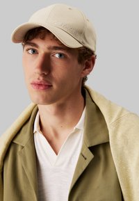Beige katoenen pet met geborduurd "Calvin Klein"-logo; gecombineerd met een olijfgroene blazer en een lichtgekleurde poloshirt. Neutraal kleurenpalet.