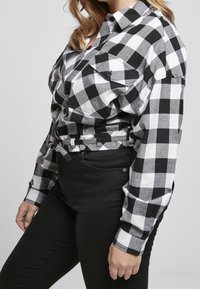 Donna che indossa una camicia a quadri bianca e nera con maniche lunghe e pantaloni neri a vita alta, su uno sfondo neutro.