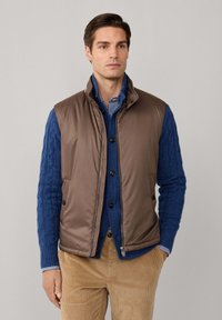 Gilet imbottito marrone indossato sopra un maglione blu a lavorazione a treccia, con colletto a scacchi. Presenta una chiusura con zip e bottoni, mostrato frontalmente.