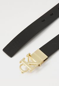 Cintura nera in pelle con texture liscia, dotata di una fibbia in metallo dorato con un logo stilizzato "CK". Regolabile con più fori.