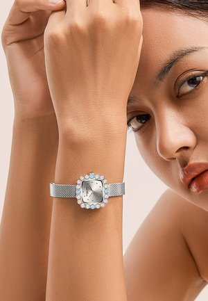 Montre à bracelet en maille argentée avec un cadran carré, entouré de pierres précieuses colorées. Elle présente un cadran minimaliste avec des marqueurs d'heure.