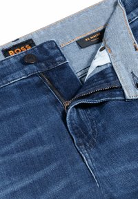 Jeans in denim blu scuro con zip e chiusura a bottone. Presentano cuciture arancioni a contrasto e etichette del marchio all'interno della cintura.