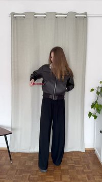 Svart bomberjakke med teksturert skinn, ribbestrikkede mansjetter og midjebånd. Løse svarte bukser. Bakgrunnen har en lys gardin og grøntområder.