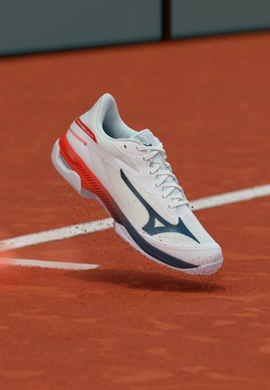 Chaussure de tennis blanche et rouge avec un logo bleu soulevant de la poussière d’argile sur un court en terre battue rouge près d’une ligne blanche.