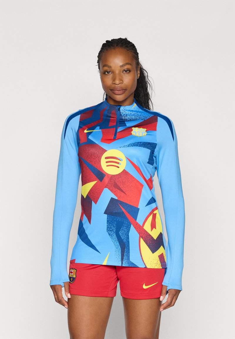 Niebieska sportowa koszulka z długim rękawem z odważnymi czerwonymi, żółtymi i niebieskimi wzorami graficznymi, noszona z czerwonymi szortami ozdobionymi logo marki.