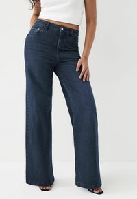 Mørkeblå brede jeans med høj talje, lavet af denim. Har fem lommer og knaplukning. Glat overflade og rene sømme.